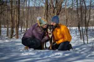 Dog mom and dog dad of an older herding dog kissing his head in the snow with woods around them