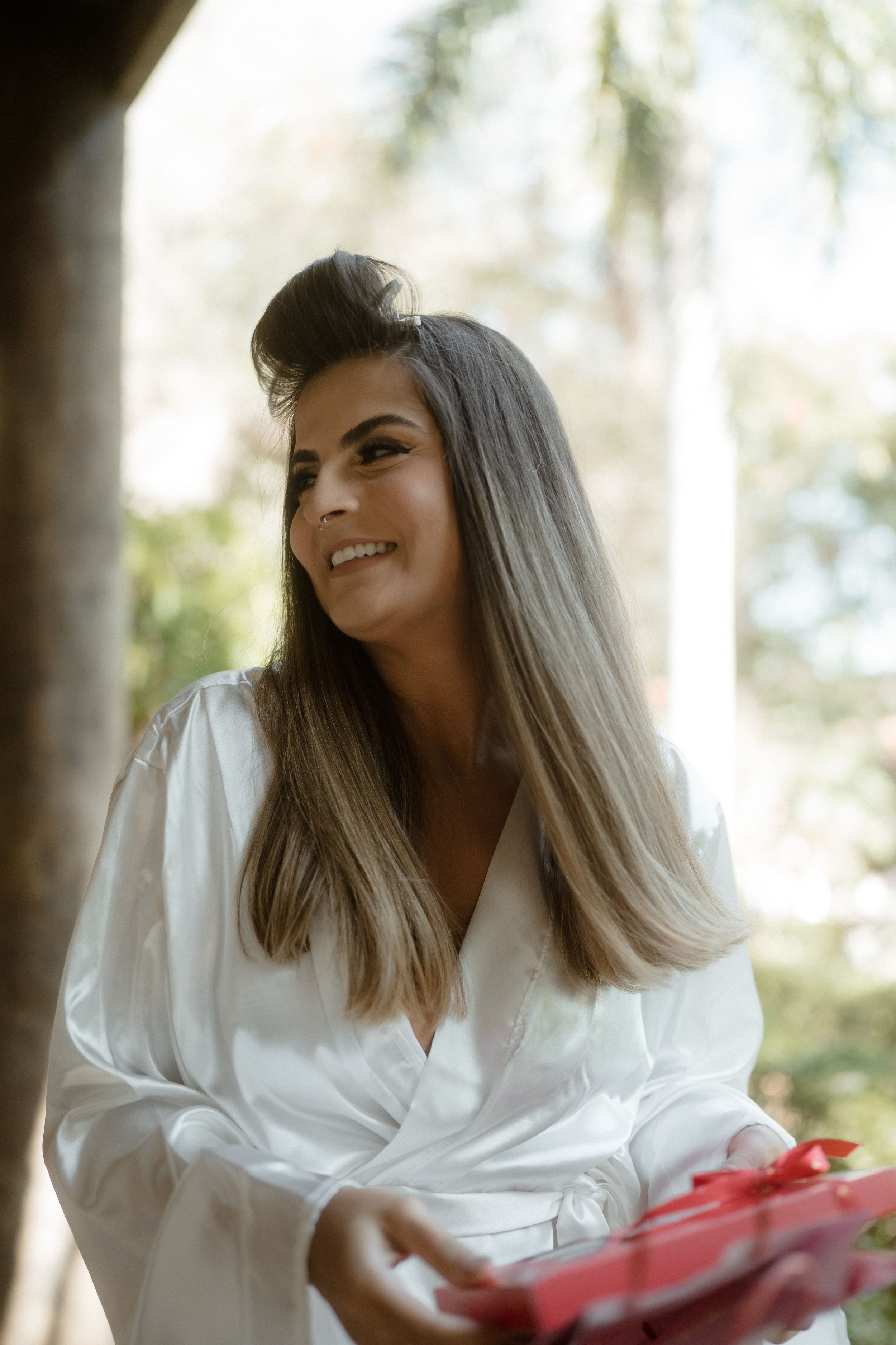 Someone wearing a white silk robe smiles while holding a red gift box outdoors.