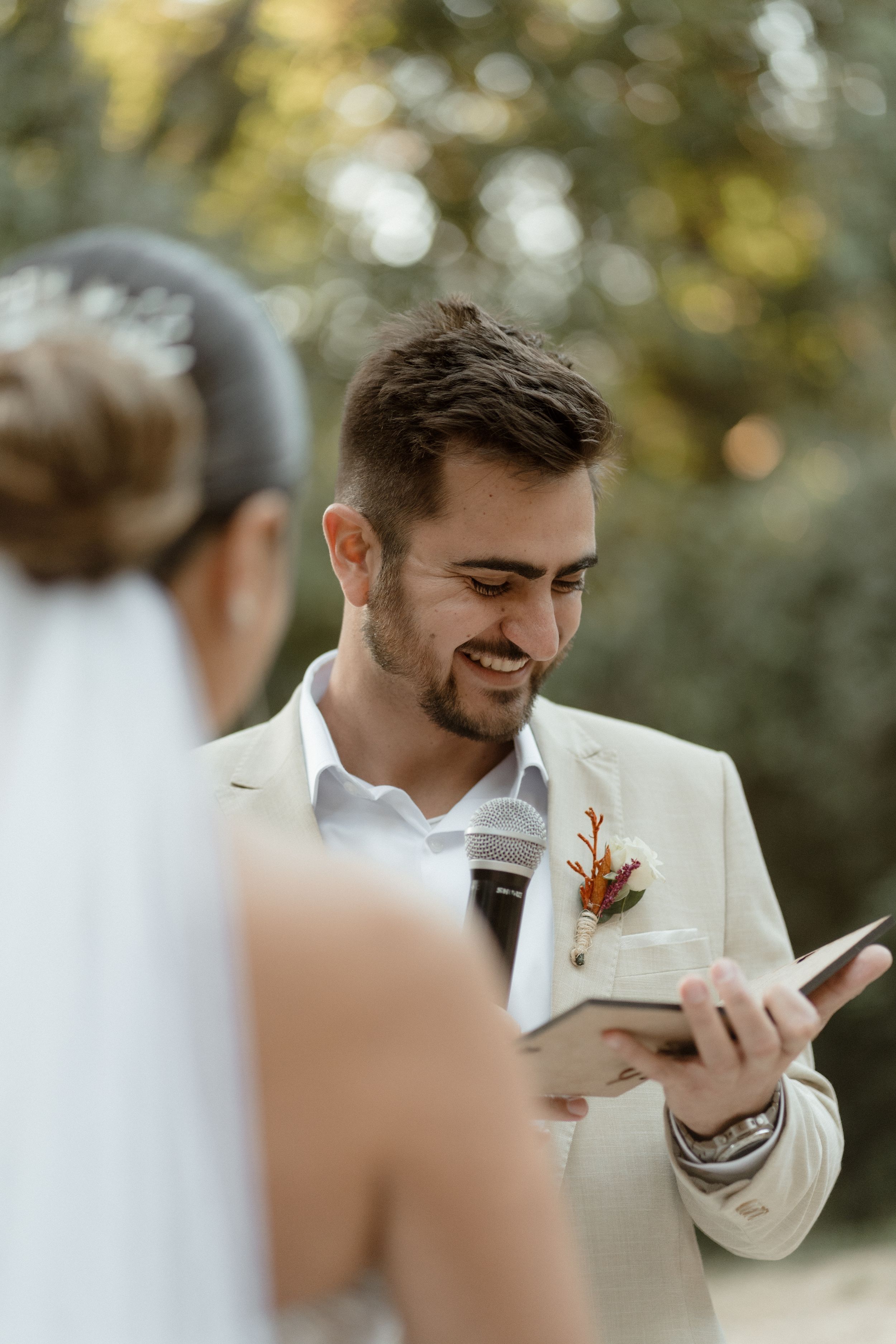 Groom in light beige suit smiling while reading from phone during outdoor wedding ceremony.