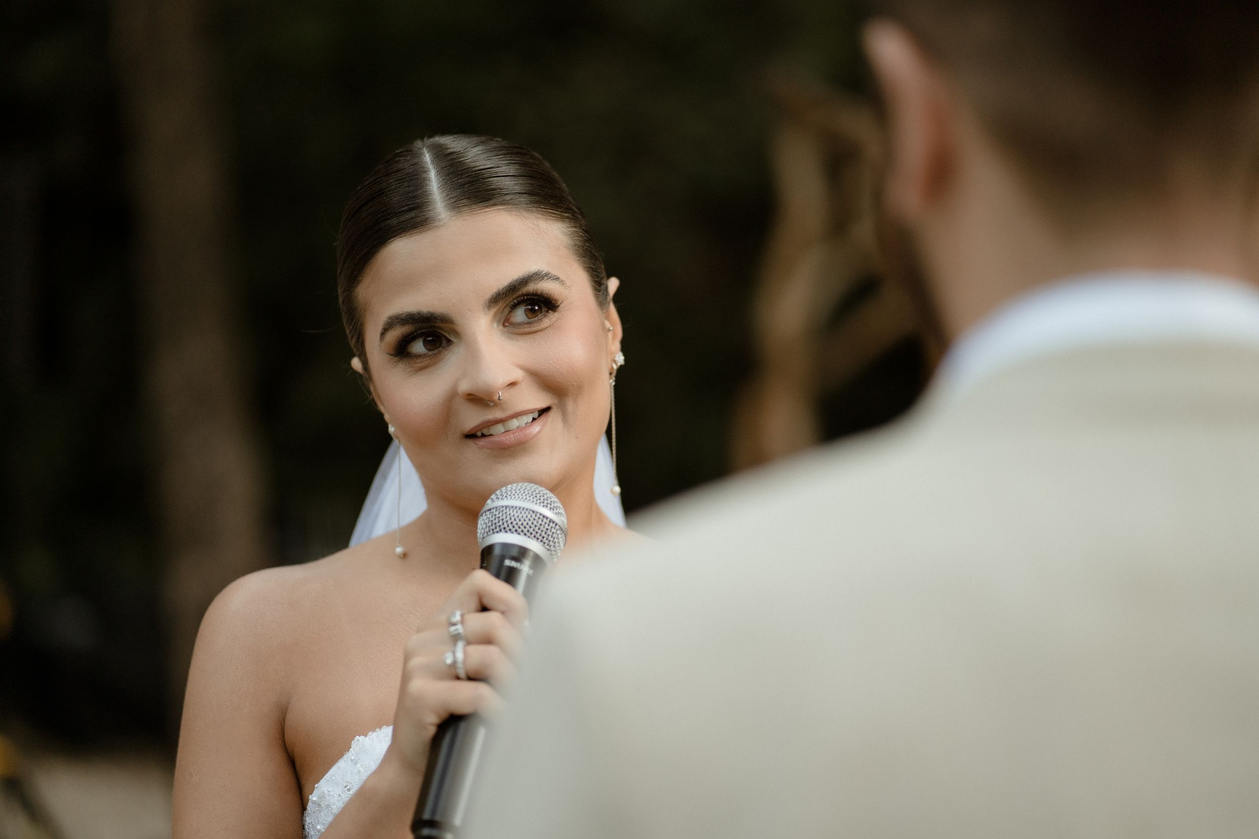 Bride speaking into microphone during outdoor wedding ceremony with natural woodland backdrop.