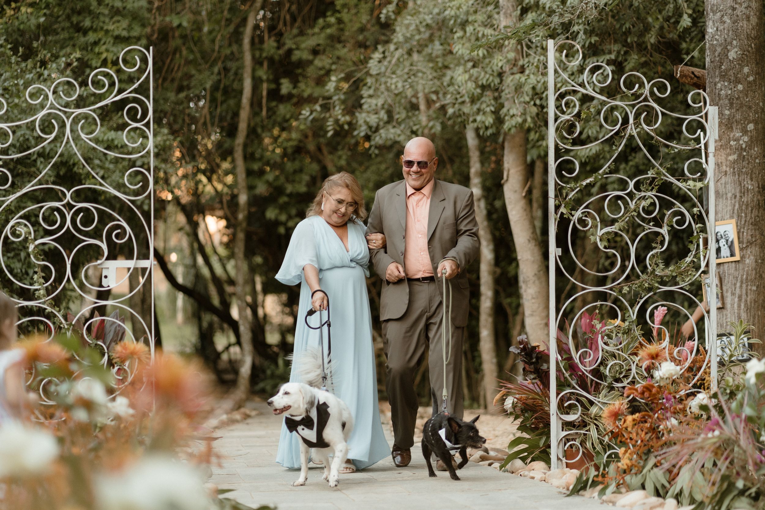 Senior couple walking through ornate garden gate with small dog during wedding celebration.