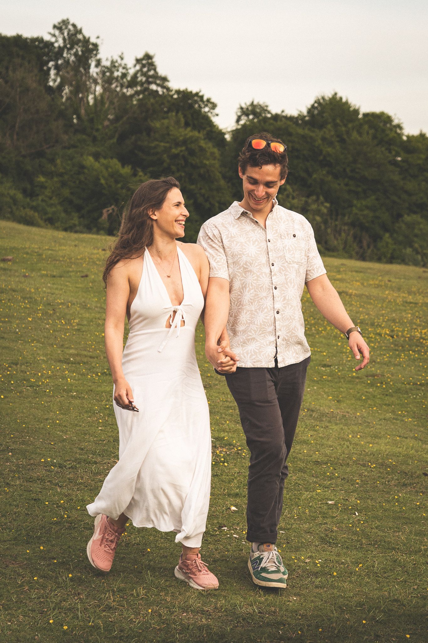 A playful couple in casual attire running and laughing together on a grassy hillside.
