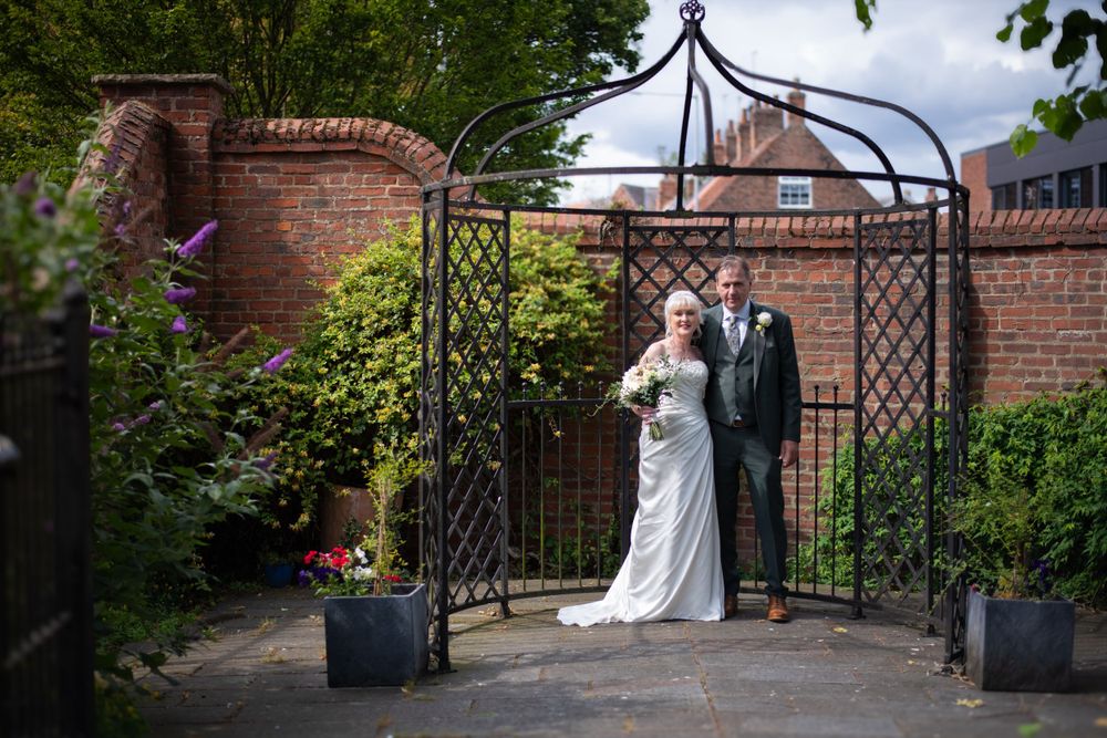 Wedding couple standing under an ornate metal garden arch with brick walls.