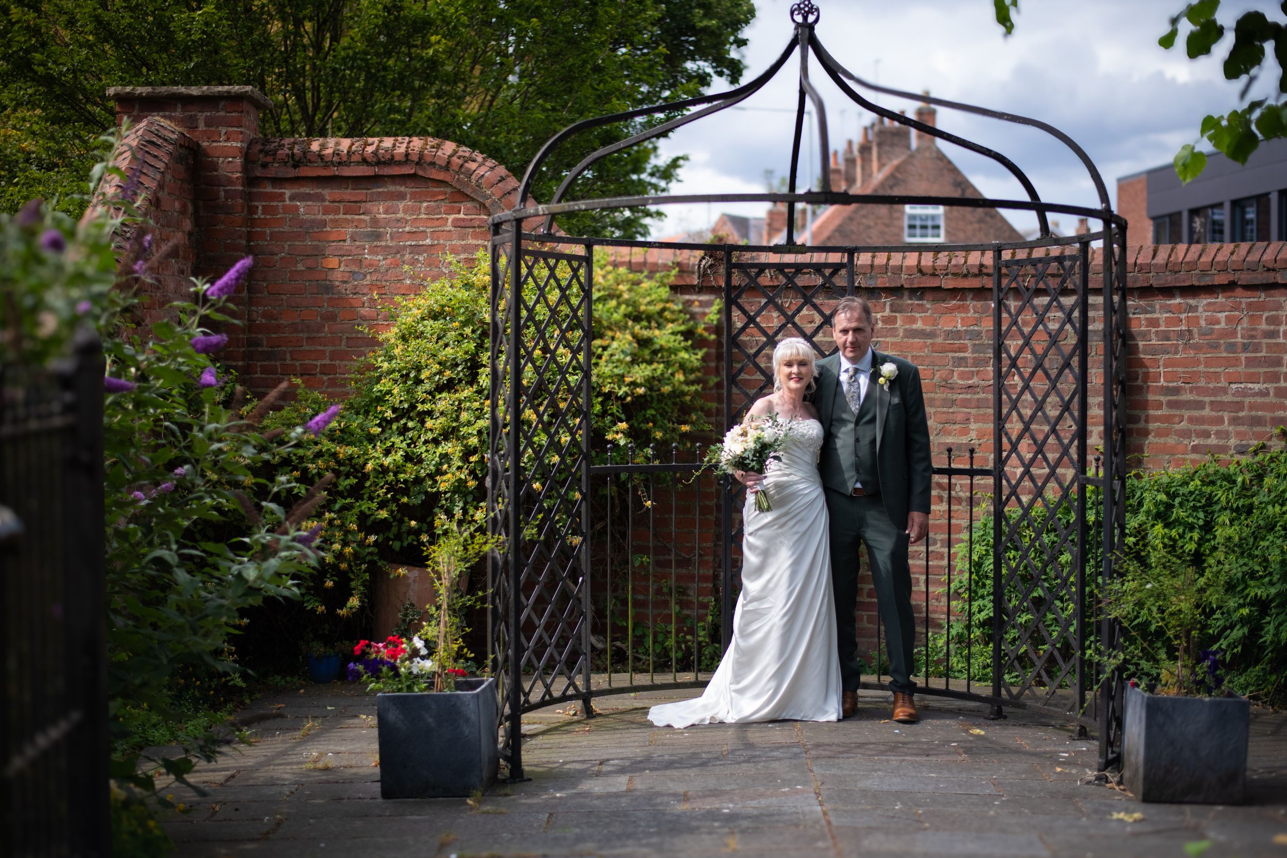 Wedding couple standing under an ornate metal garden arch with brick walls.