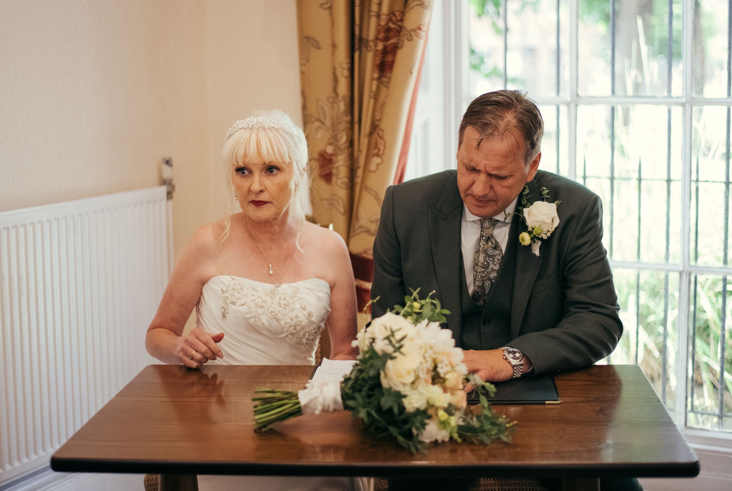 A couple signing marriage documents at a small desk by a window.