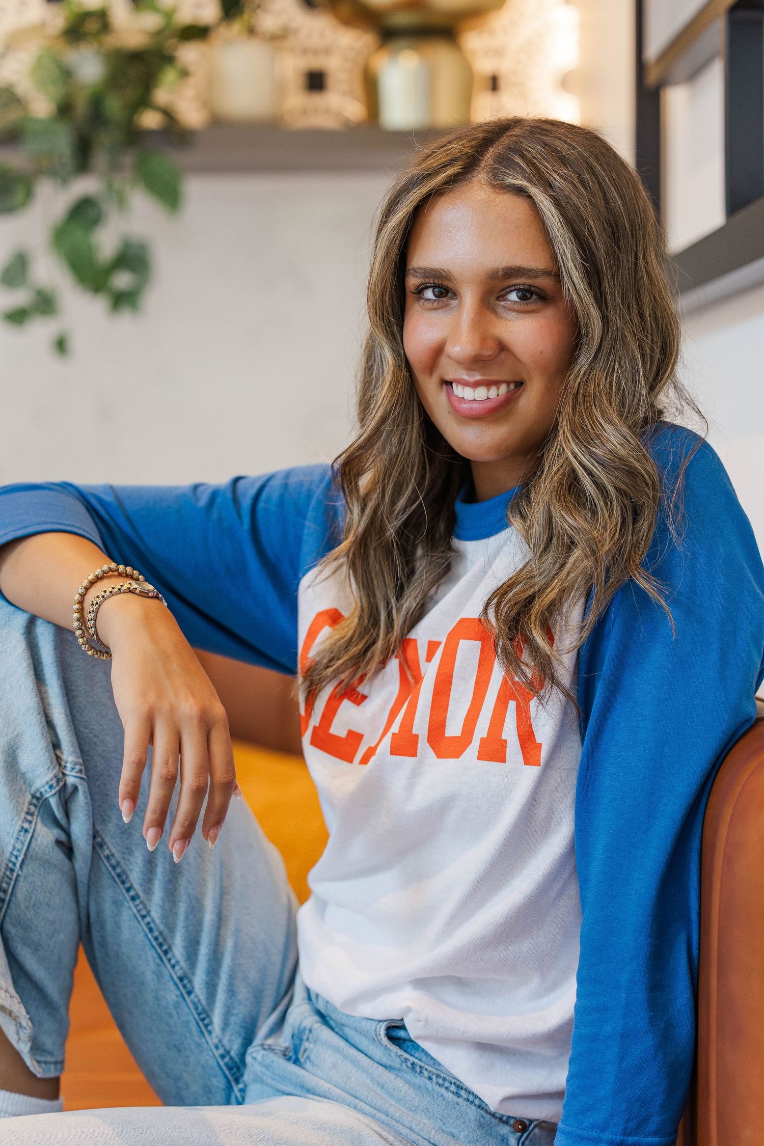 Smiling young woman in blue raglan senior shirt sits relaxed indoors with wavy highlighted hair and jeans.