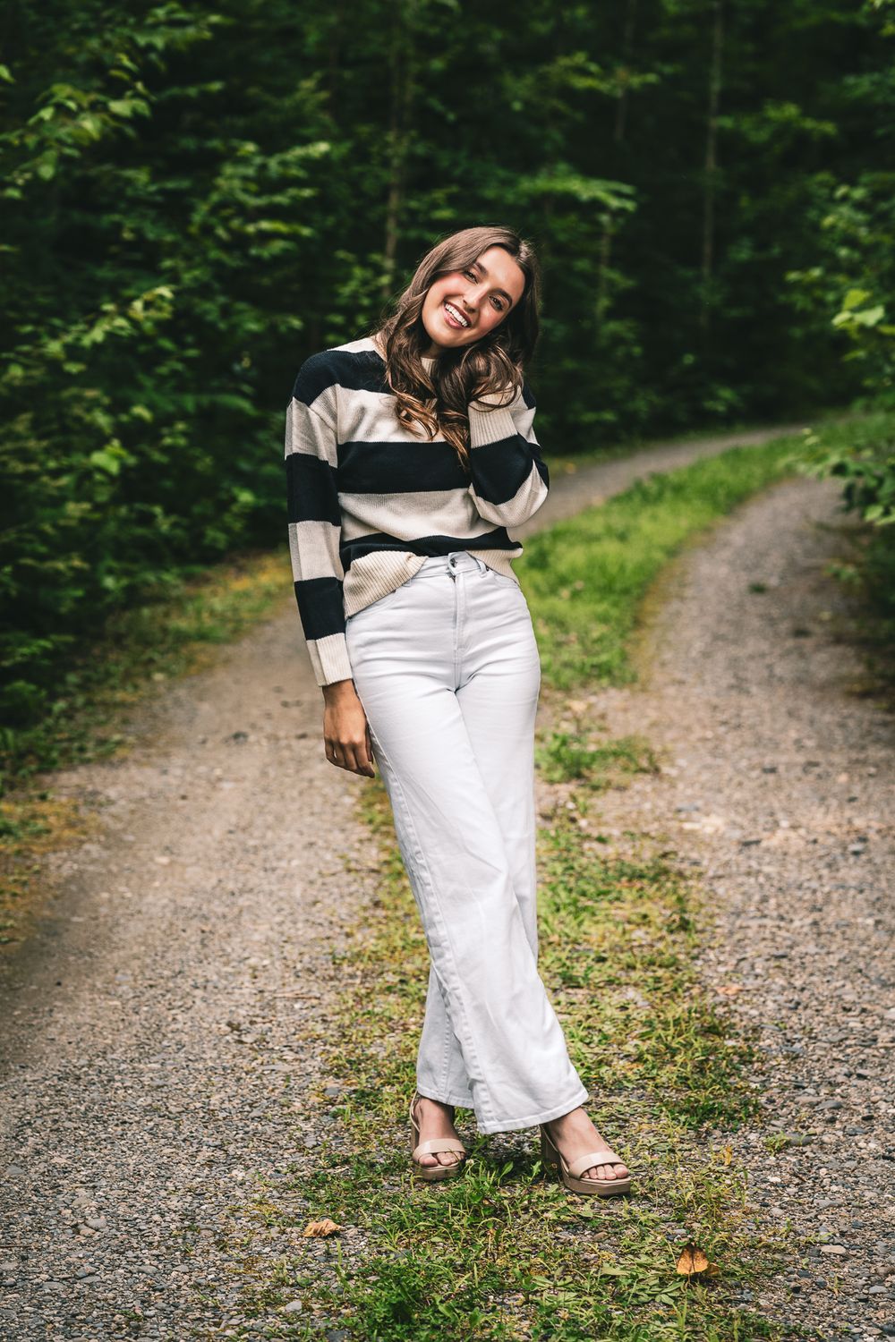 Woman in striped sweater and white pants posing casually on forest trail.