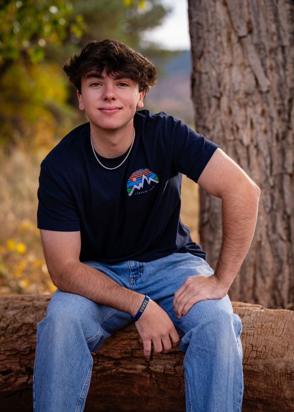 Senior portrait of teen in navy t-shirt sitting casually on wooden log outdoors.