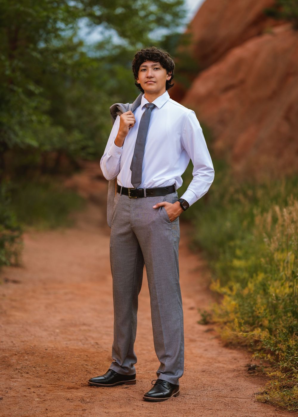 Professionally dressed person in business attire stands on red dirt path with rock formations in the background.