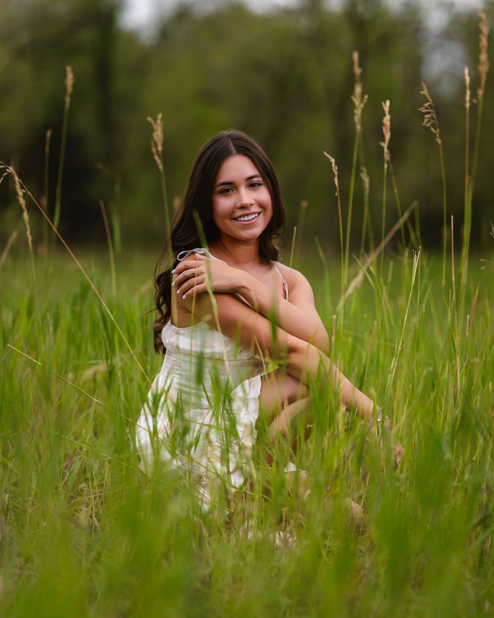 A person in a white dress crouches among tall green grass stalks in an outdoor field setting.