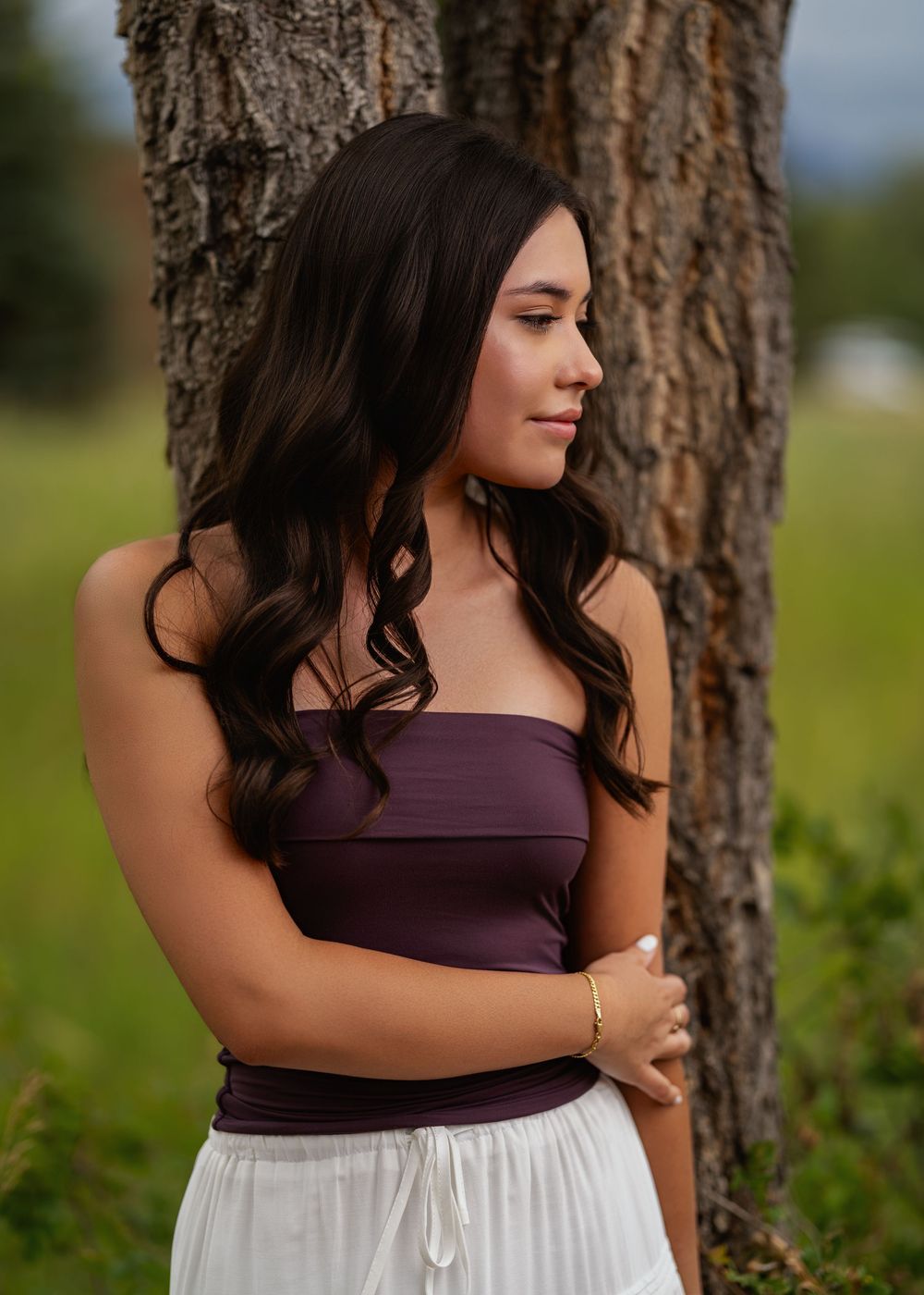 Profile view of someone in a purple strapless top and white flowy skirt standing against a tree trunk outdoors.