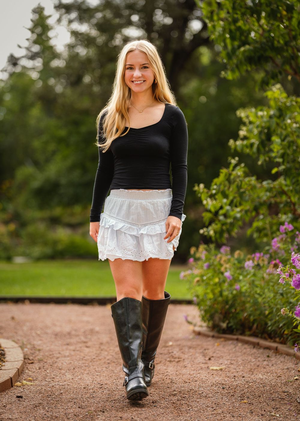 A person wearing a black top and white lace skirt strolls along a gravel garden path bordered by bushes and flowers.