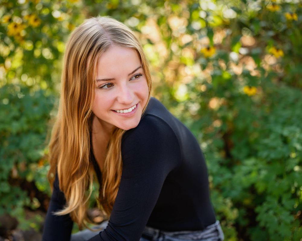 A person wearing a black sweater smiles warmly while posing outdoors with autumn foliage in the background.