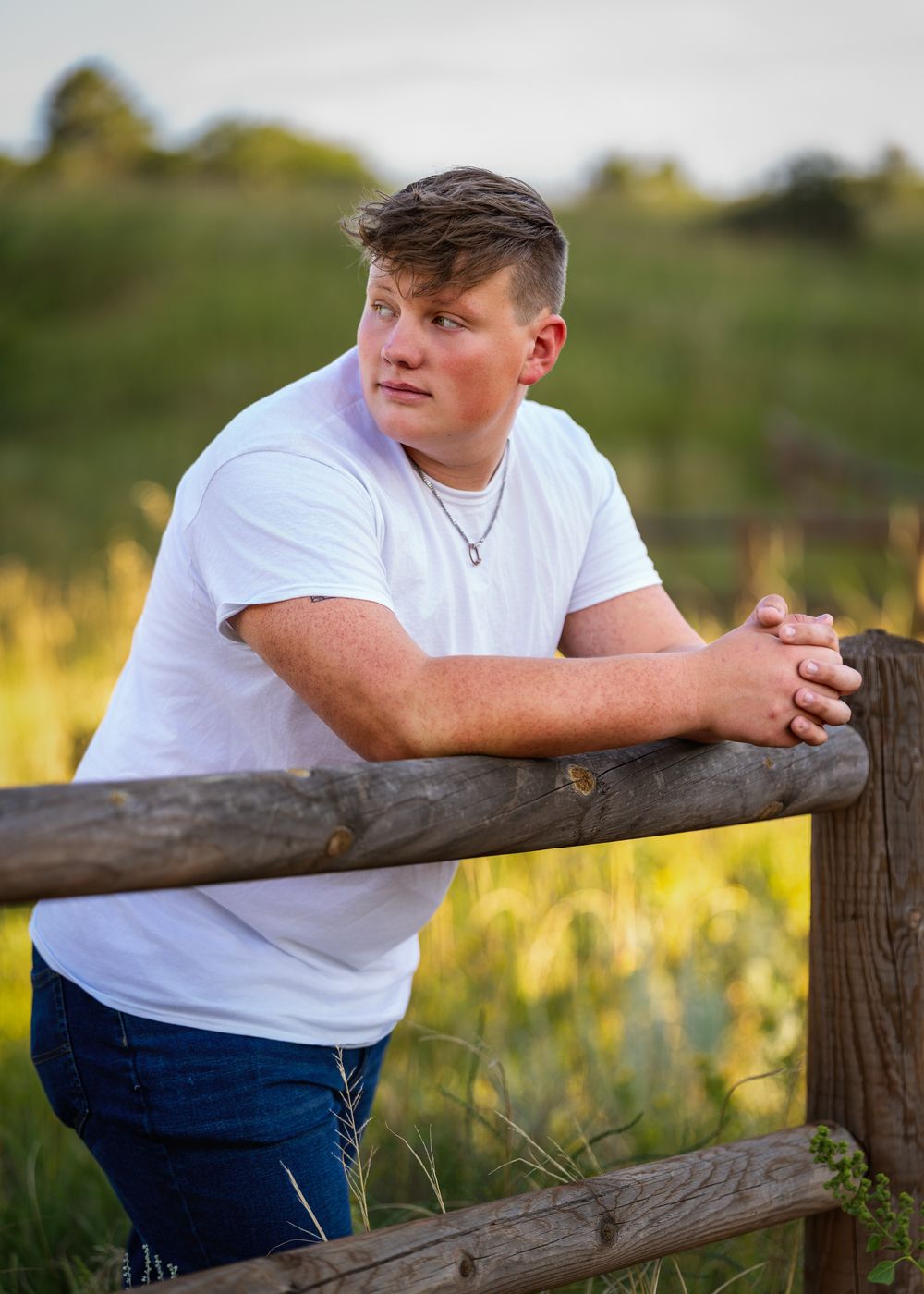 Young man in white t-shirt leaning on rustic wooden fence in a green meadow during golden hour.