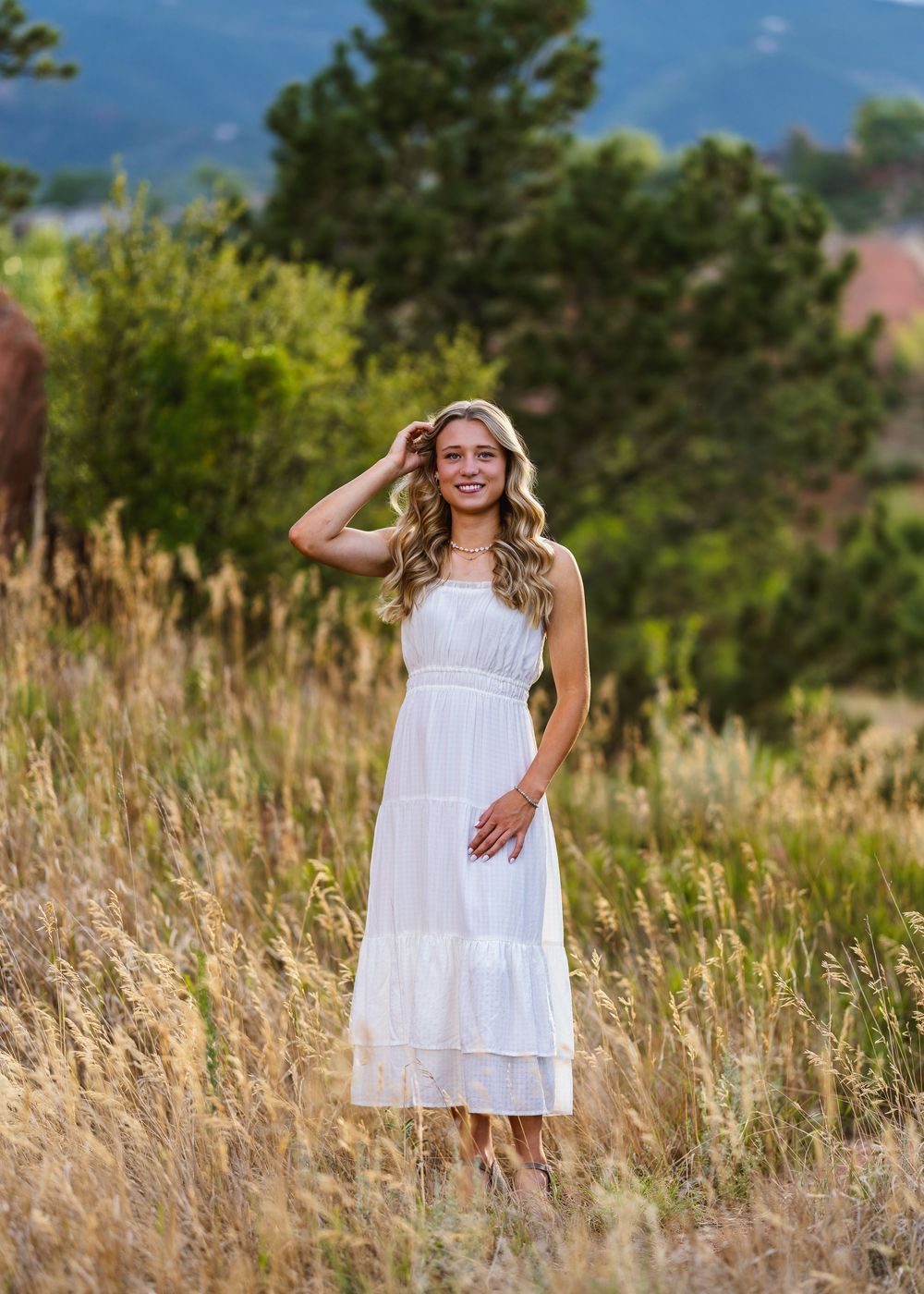 Young woman in white midi dress standing in golden field with mountain backdrop on sunny day.