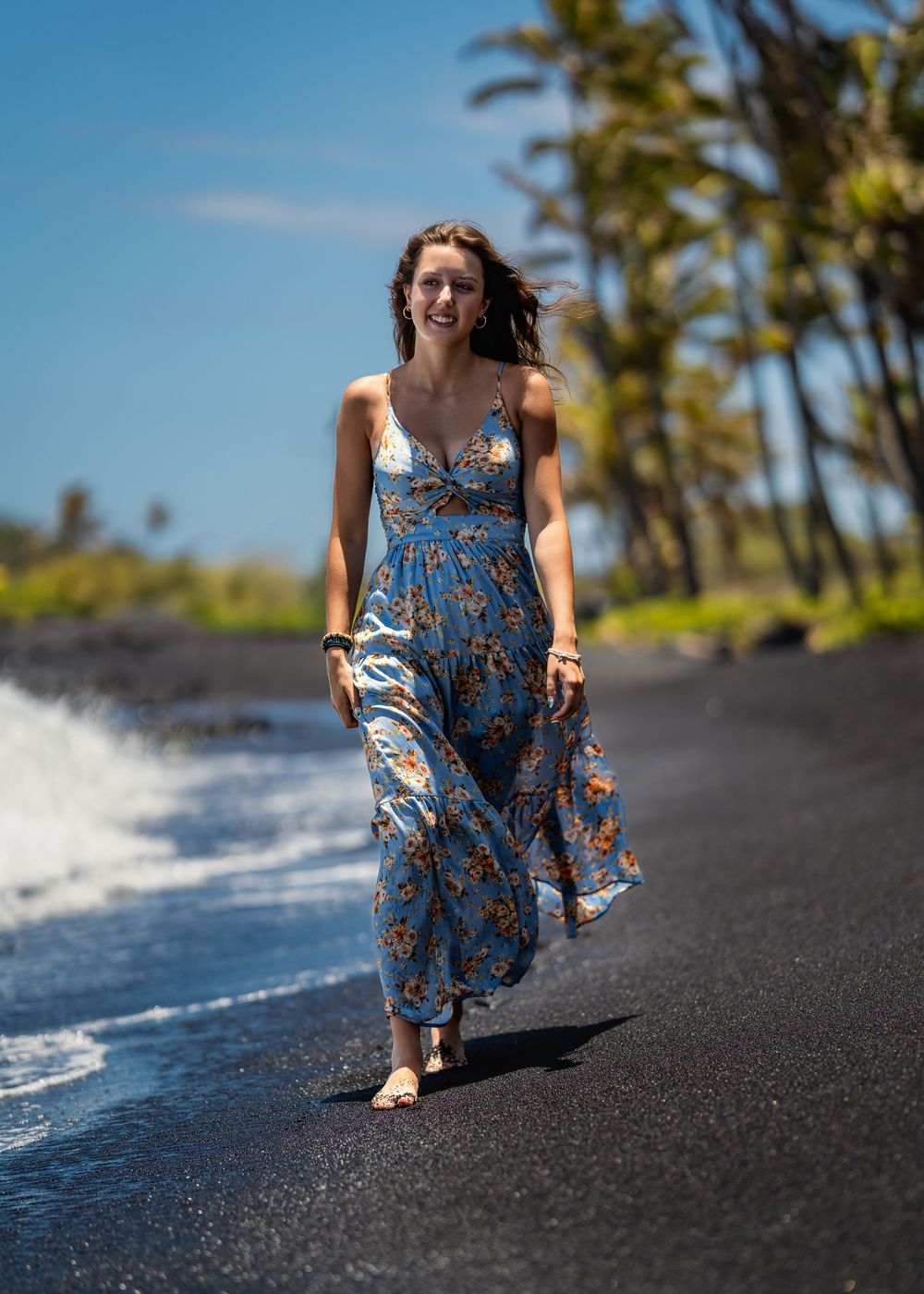 A flowing blue floral dress moves gracefully against black sand and ocean waves at a Hawaiian beach.