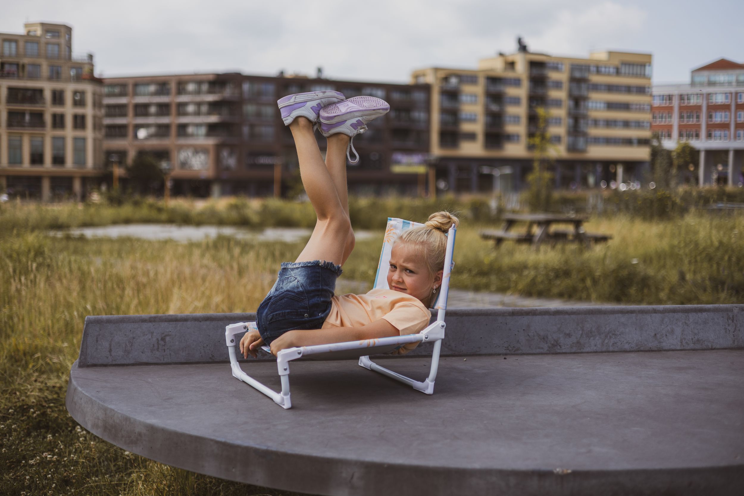 A baby relaxes on a tiny lounge chair outdoors with legs up in the air, set against an urban backdrop.