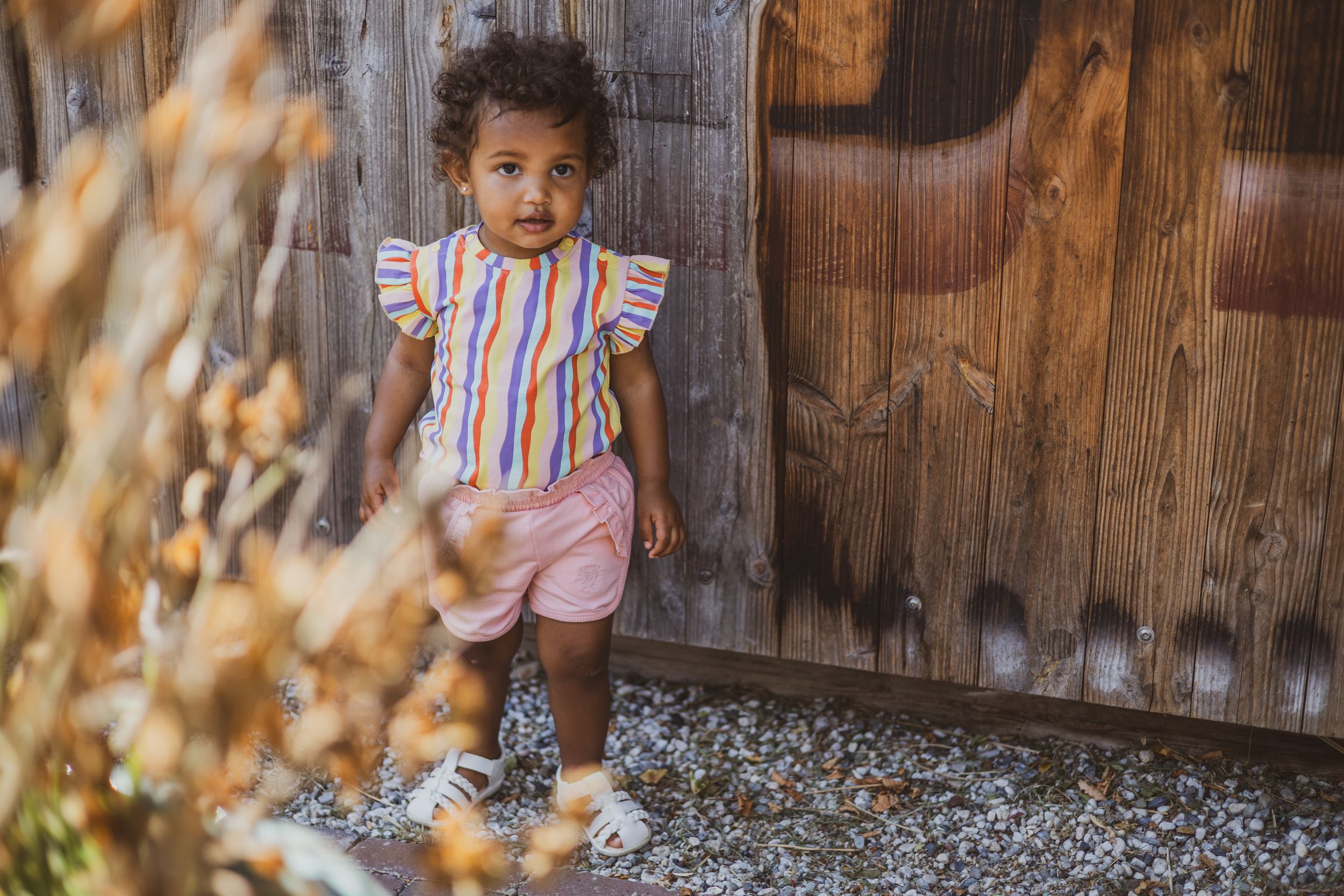 Toddler girl in colorful striped top and pink shorts standing by rustic wooden fence outdoors.