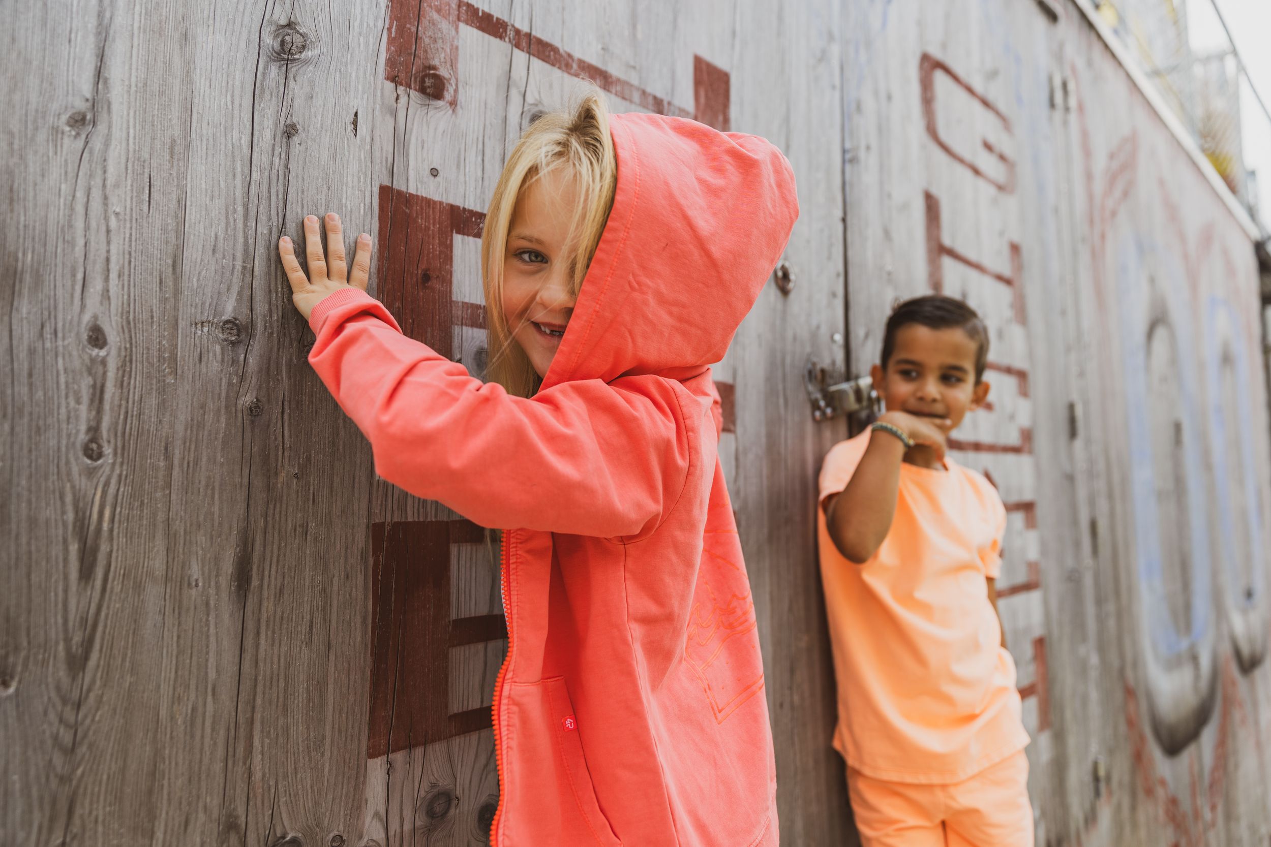 Two children in coral hoodies playing against a rustic wooden wall outdoors.