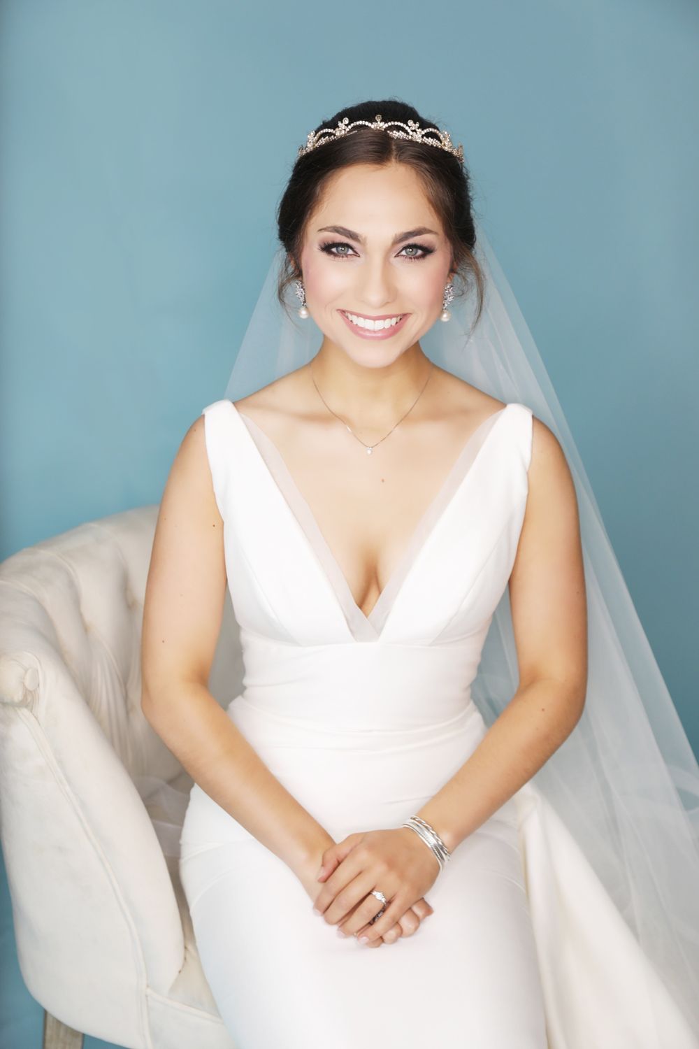 A bride wearing a v-neck wedding dress and tiara smiles against a blue background.