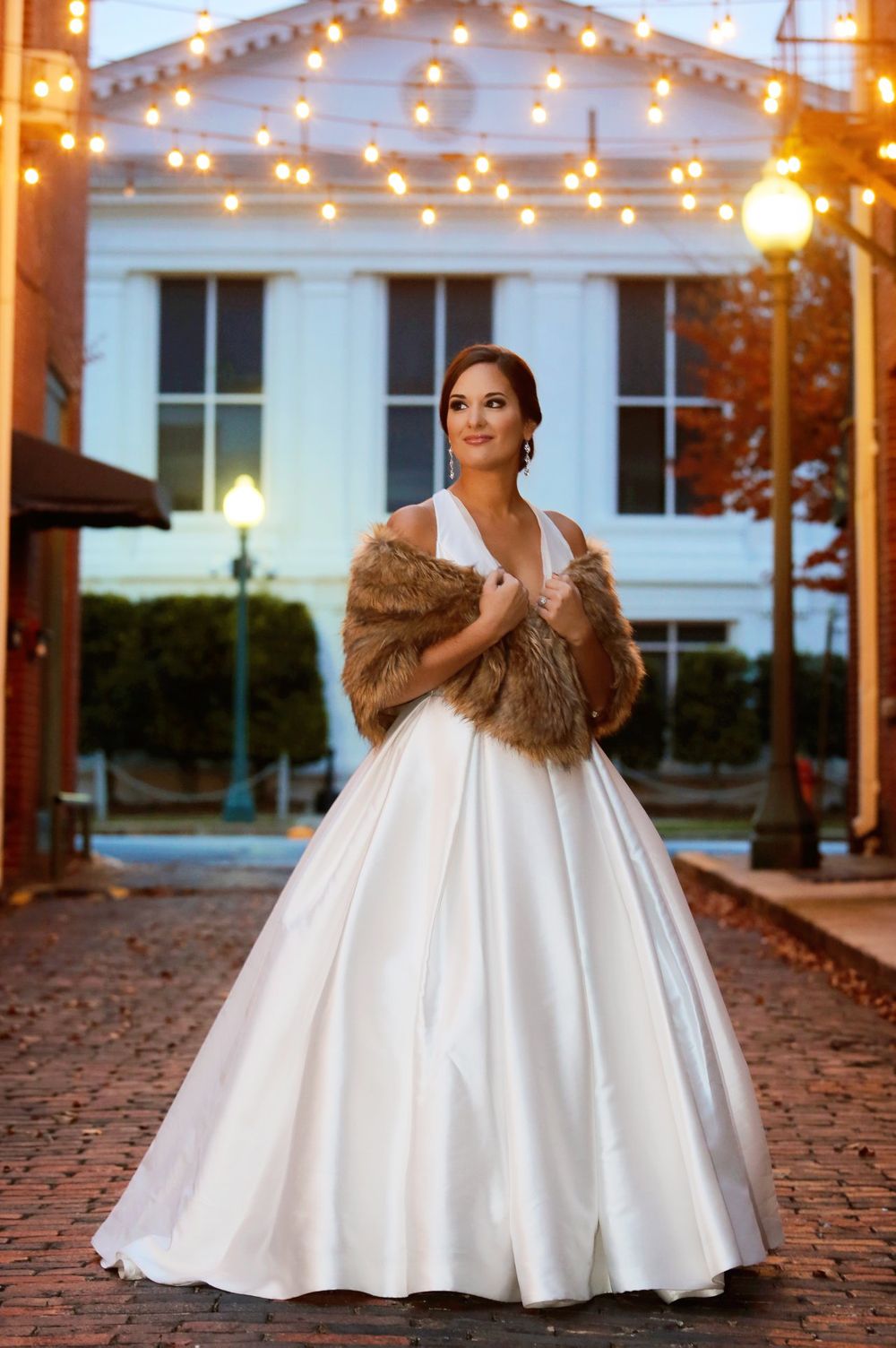 Bride in classic white ballgown with fur stole stands under romantic string lights at dusk.