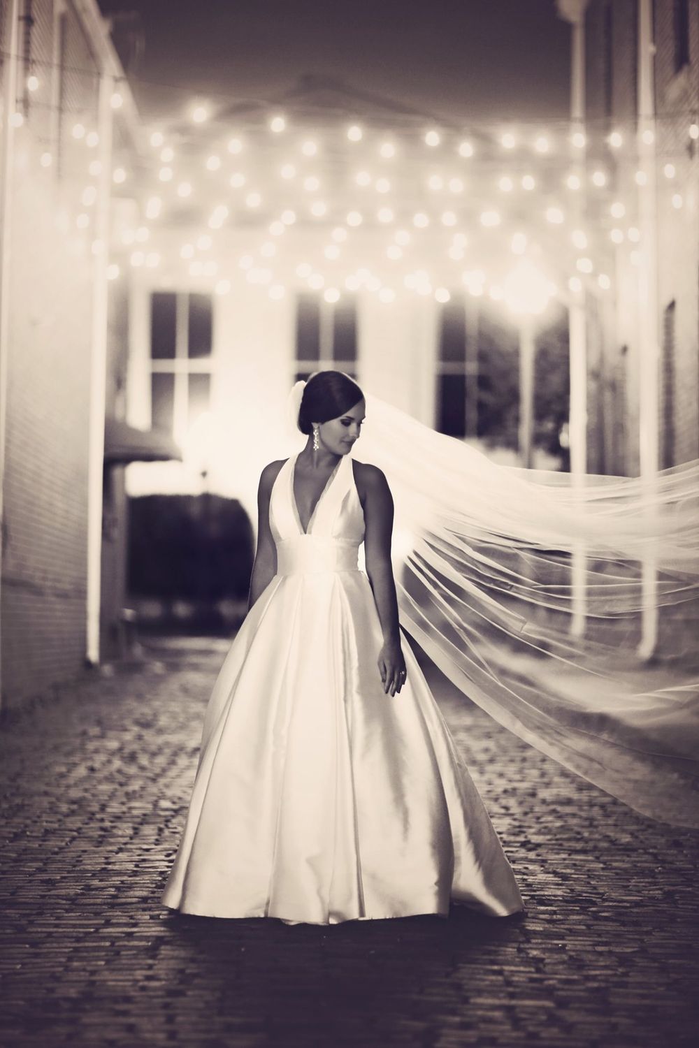 Dramatic black and white bridal portrait under string lights on cobblestone path.