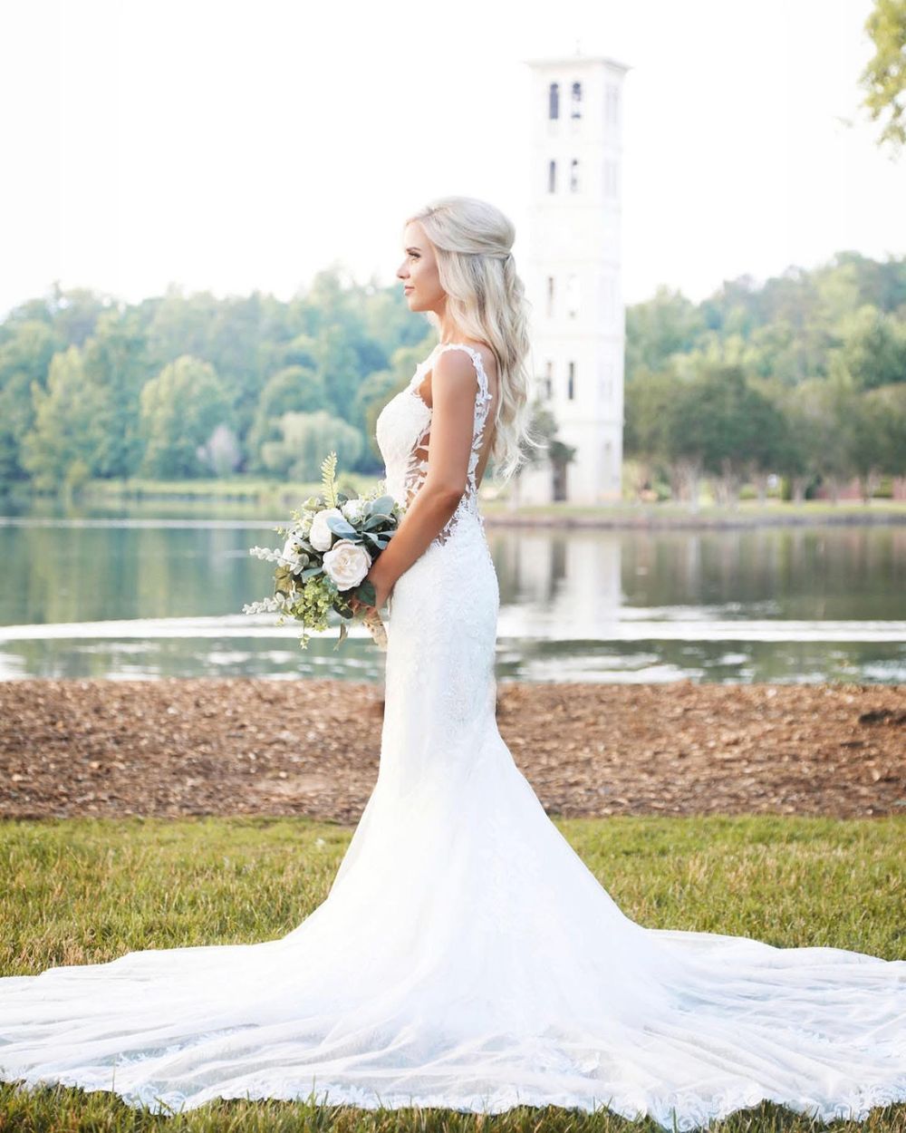 A bride in a flowing white wedding dress poses gracefully against a lakeside backdrop with a white bouquet.