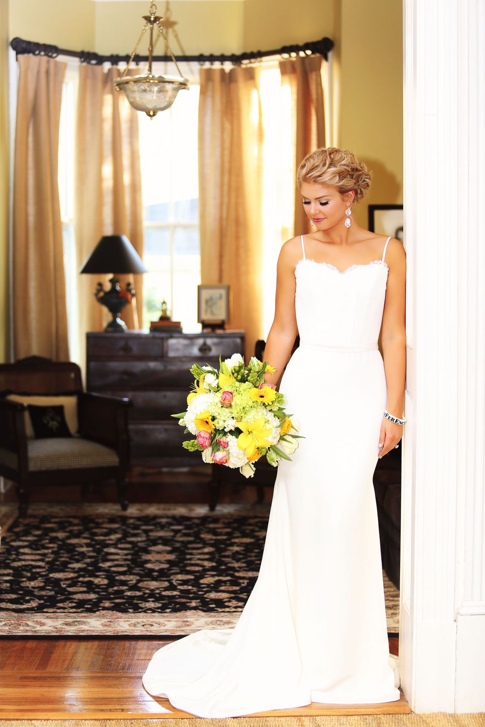 A bride in an elegant sheath dress holds a yellow bouquet in an ornate interior setting.