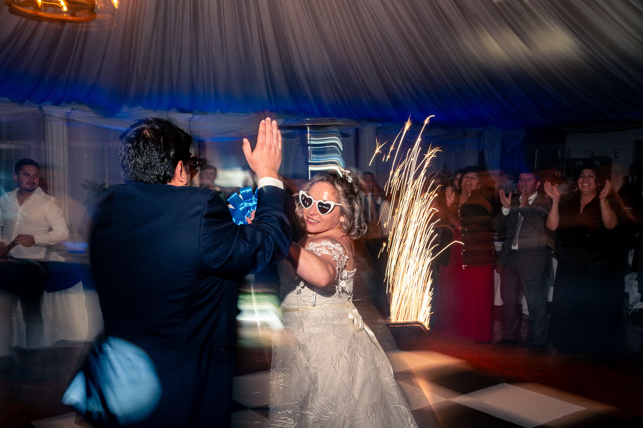 Parejas bailando en recepción de matrimonio con efectos de luz de bengalas sobre pista de baile.
