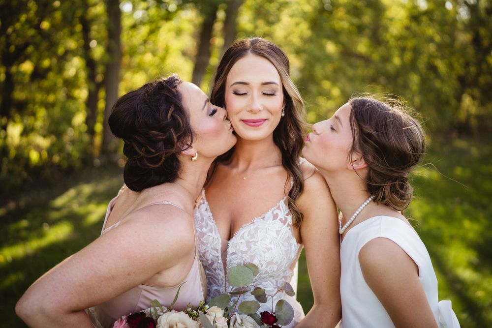Bridesmaids in blush dresses share affectionate moment with bride outdoors.