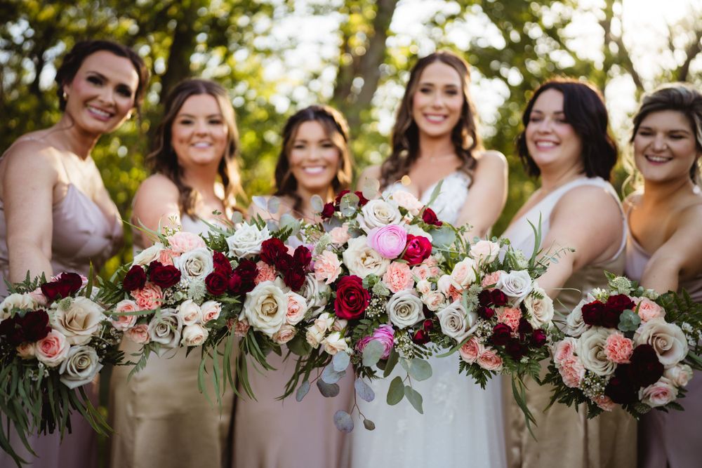 Bridal party showcases elegant bouquets of pink and burgundy flowers in outdoor setting.