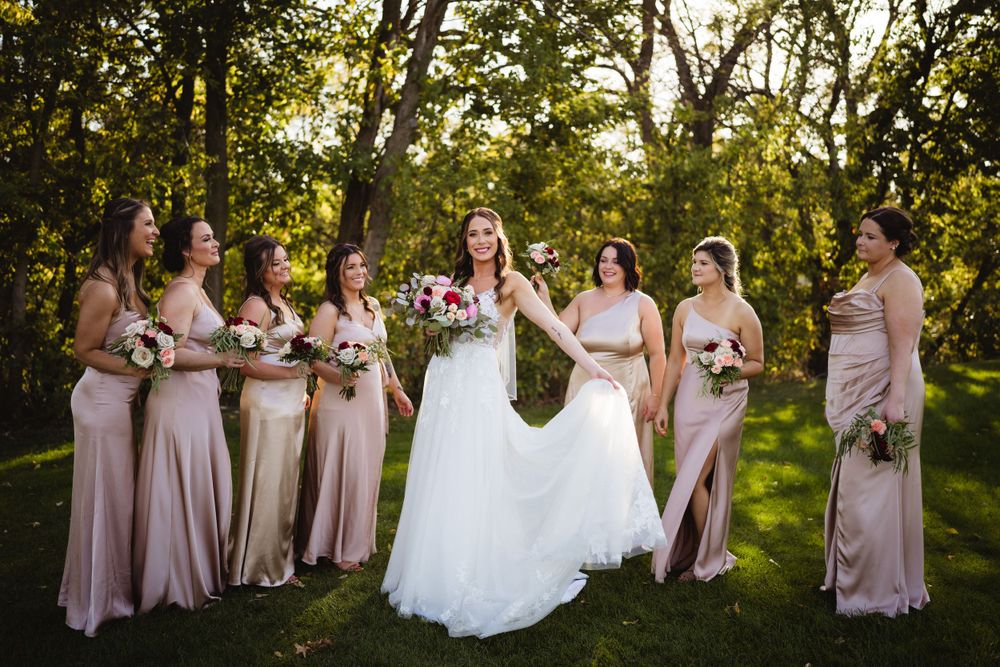 Bridesmaids in blush pink dresses stand with bride in white gown for outdoor wedding portrait in wooded setting.