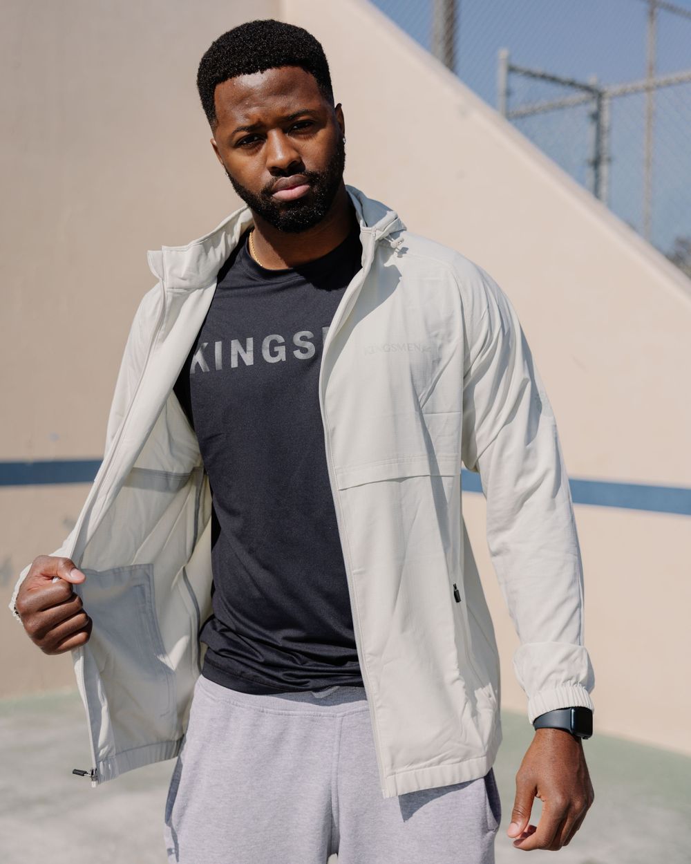 Athletic man in light jacket, dark t-shirt with KINGS text and light sweatpants stands against beige wall outdoors.