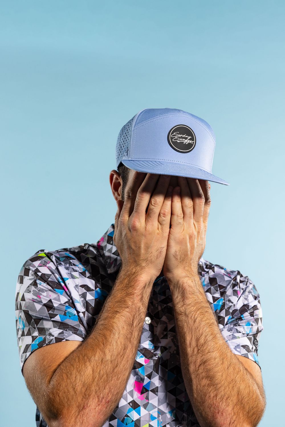 Person in geometric patterned shirt and baseball cap covers face with hands against light blue background.