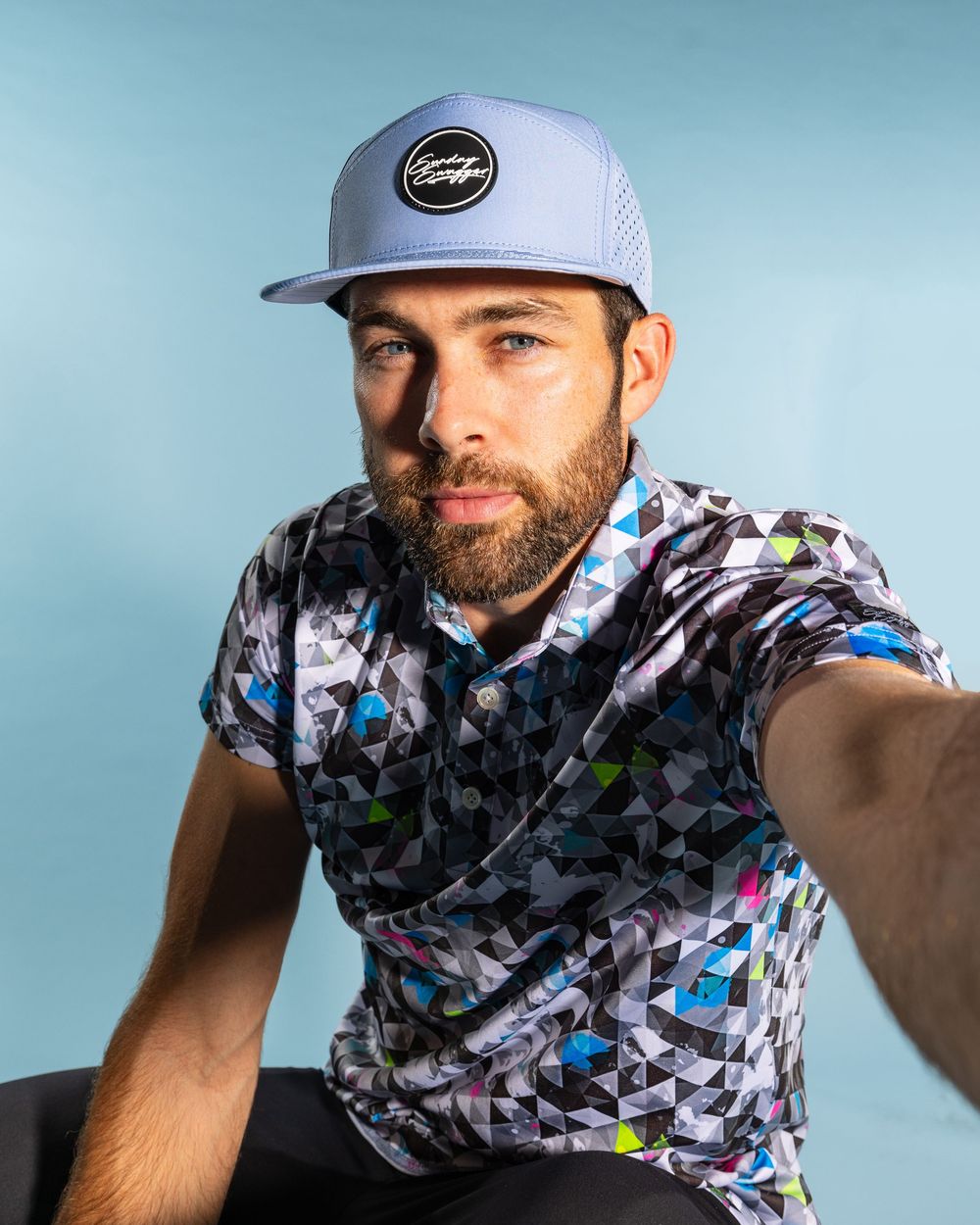 Athletic figure wearing a gray baseball cap and colorful geometric patterned polo shirt poses against blue backdrop.