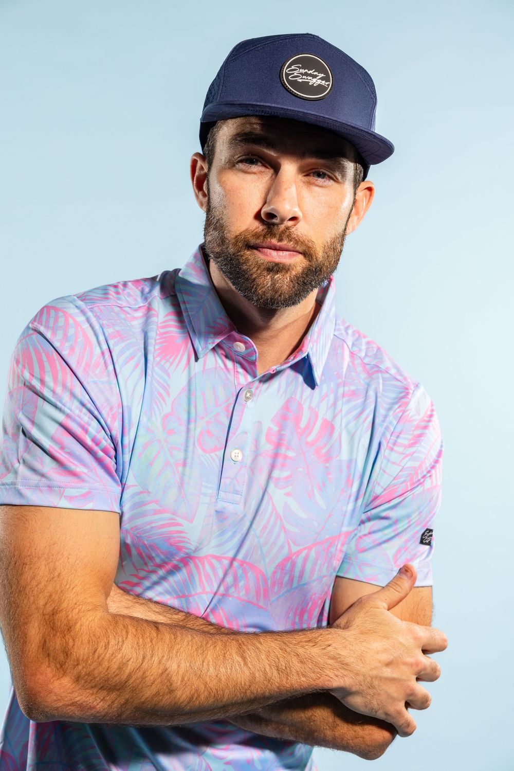 Athletic golfer wearing navy cap and pink-blue patterned polo shirt posing against blue sky background.
