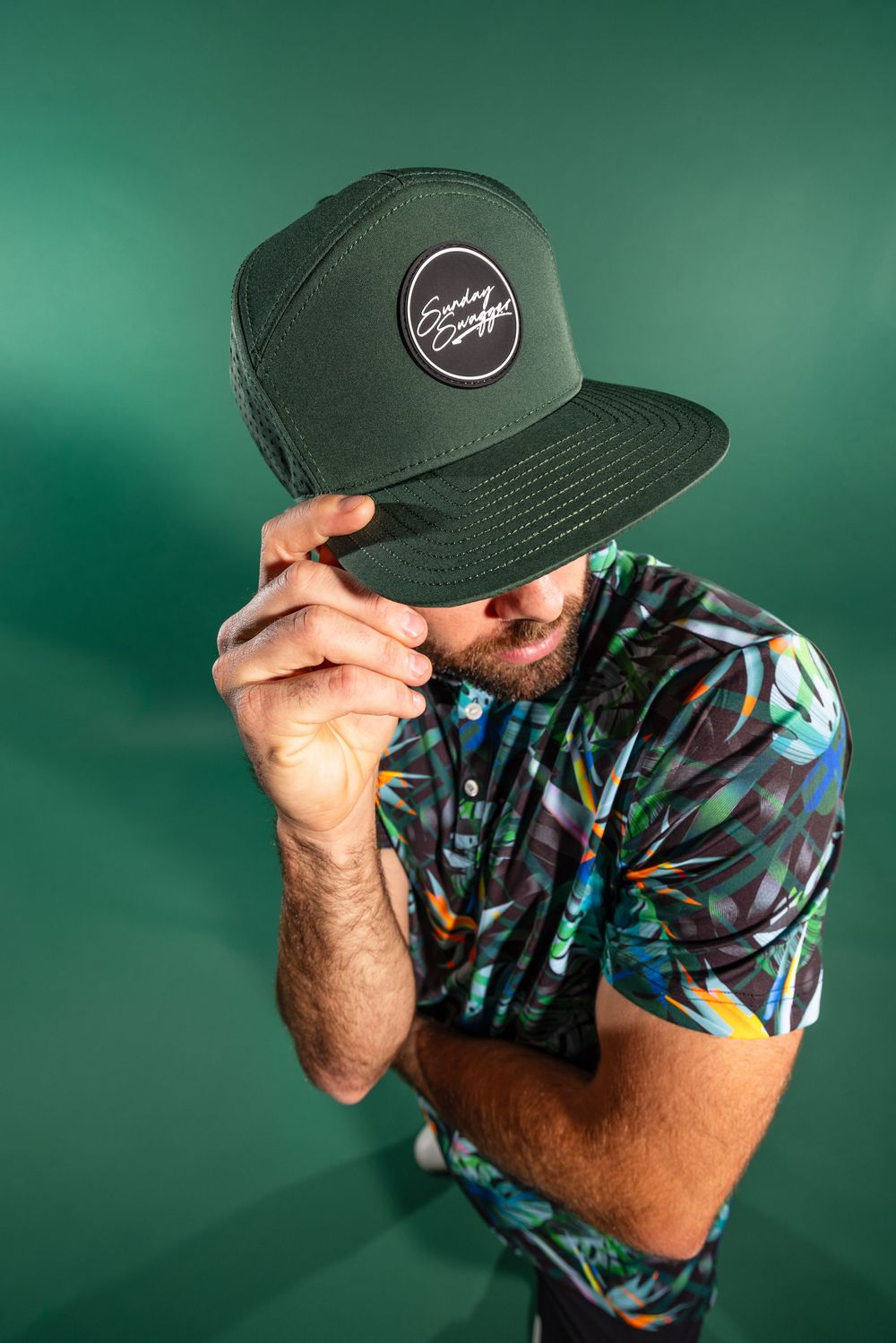 Someone in a tropical print shirt tilts their head down while adjusting the brim of a dark snapback cap against a green background.
