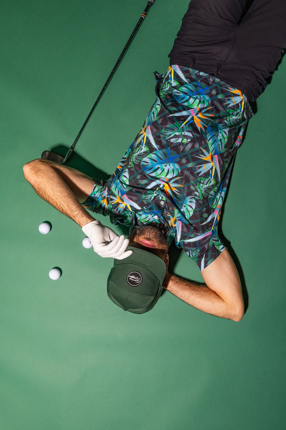 Overhead view of golfer wearing tropical print shorts lying on green putting surface with golf balls and putter.