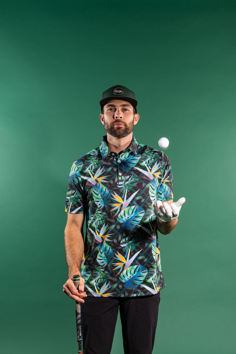 Professional golfer wearing floral Hawaiian print polo shirt tosses golf ball while posing against green background.