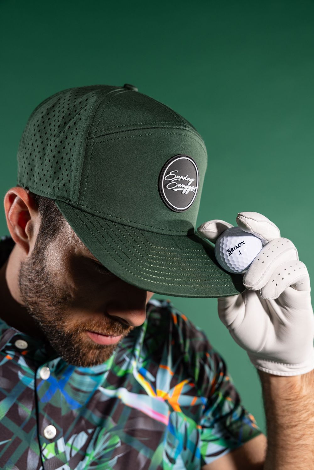 Bearded golfer in dark green cap and colorful shirt holding golf ball while looking down.