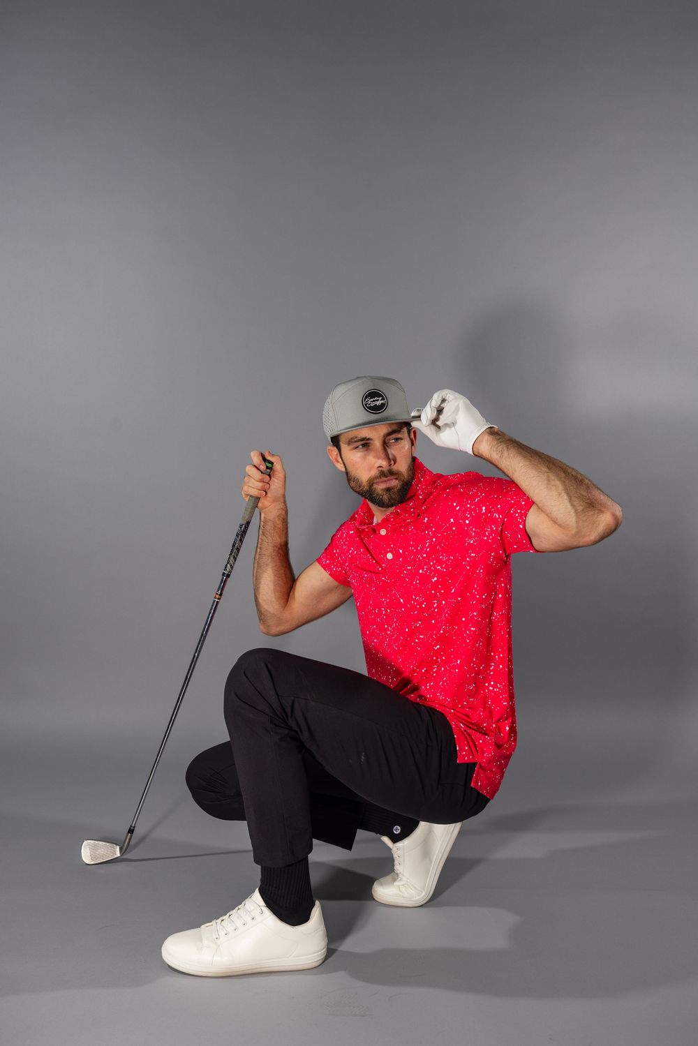 Golfer in red polka dot shirt and black pants crouches with club while posing in studio against gray background.