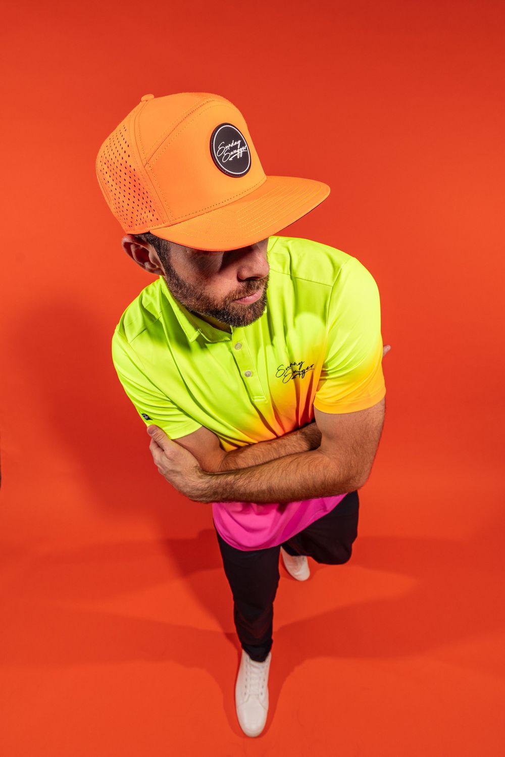 Figure wearing bright neon yellow and pink gradient shirt with orange cap stands against red background with arms crossed.