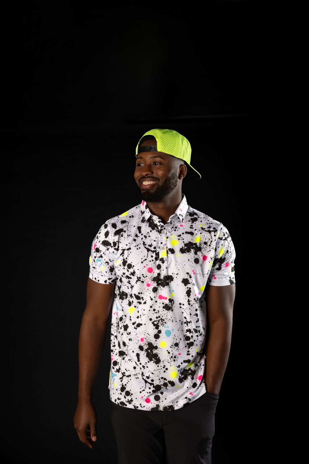A person wearing a neon yellow baseball cap and splatter-painted white polo shirt poses against a black background.