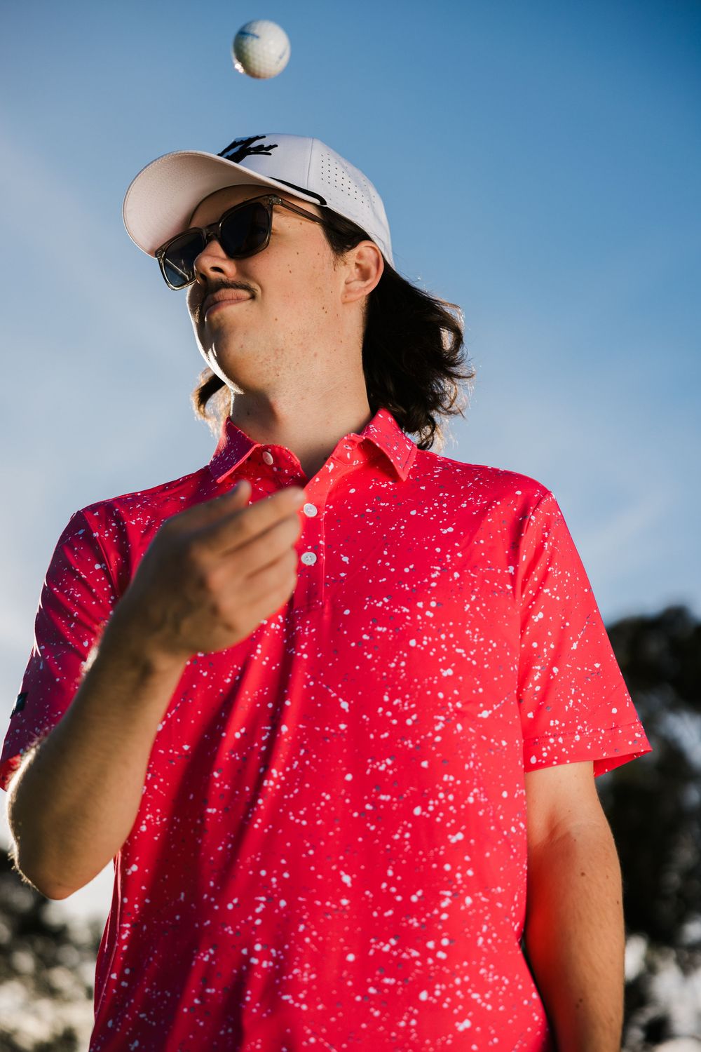 A golfer in a red speckled polo shirt and white cap throws a golf ball into the air against a blue sky background.