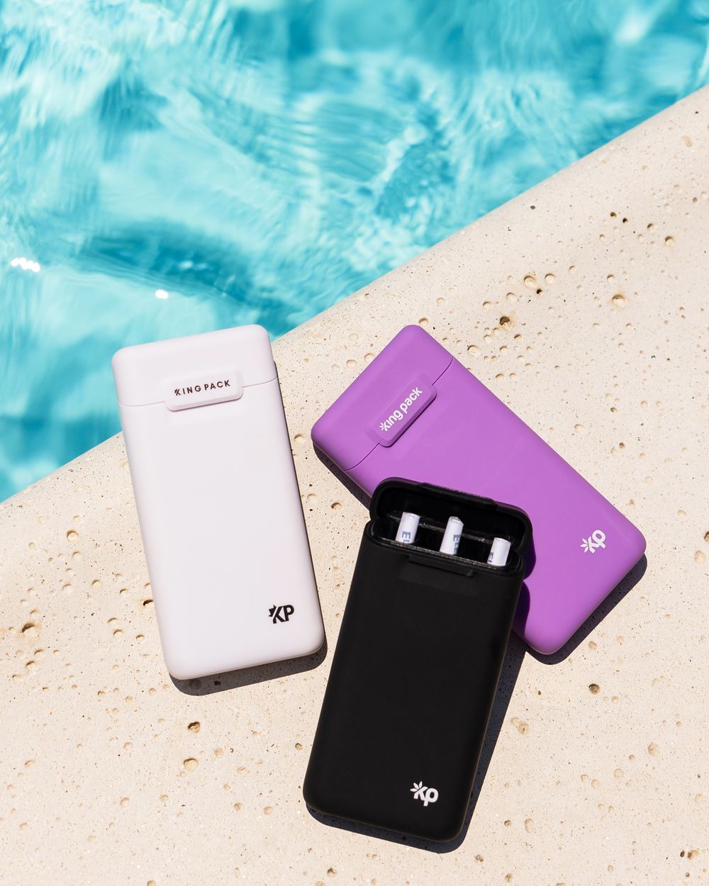 Three vape device charging cases in white, purple, and black displayed against a poolside background with blue water.