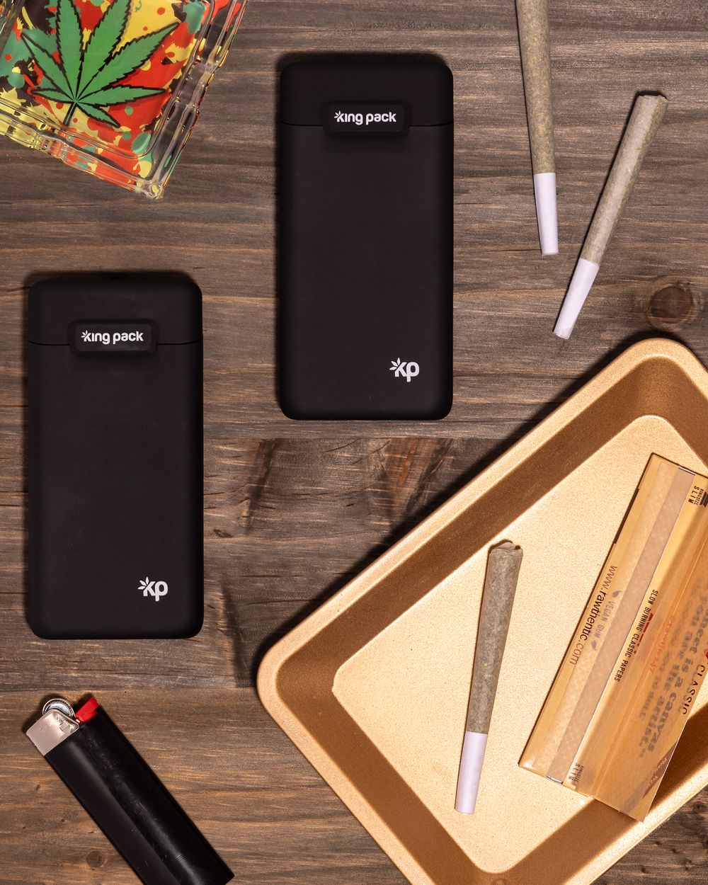 Black vape charging cases and accessories arranged on wooden surface with rolling papers and tray.