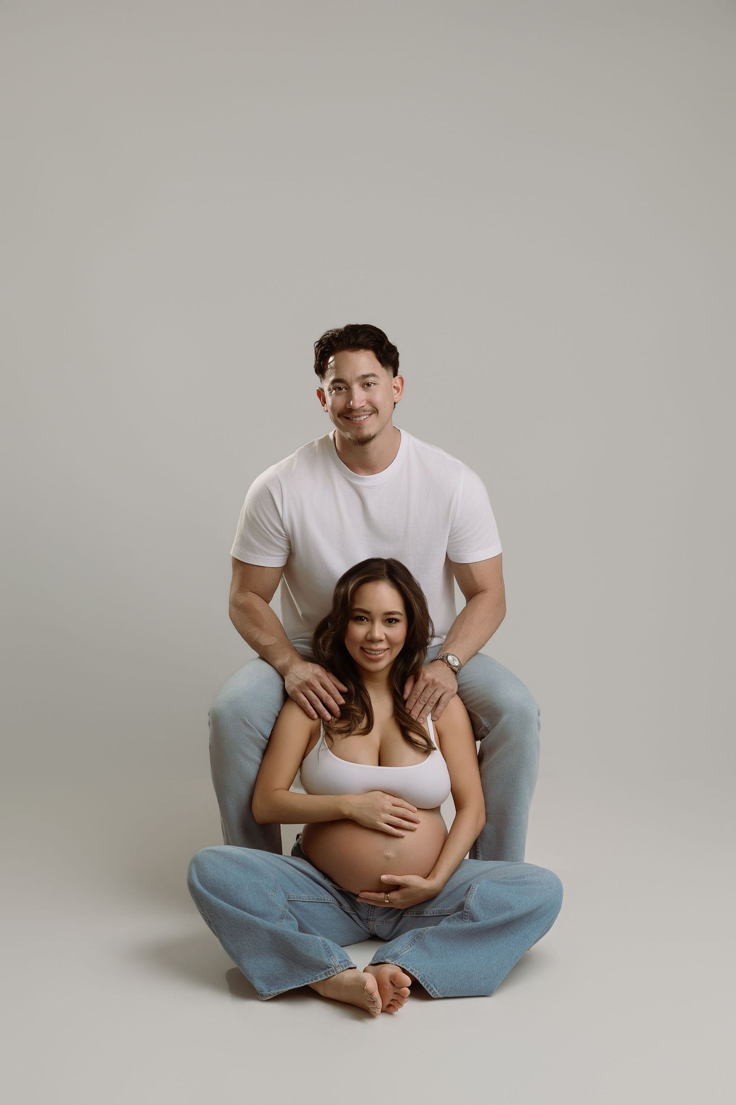 A couple sits together in a minimalist studio setting wearing white tops and blue jeans.