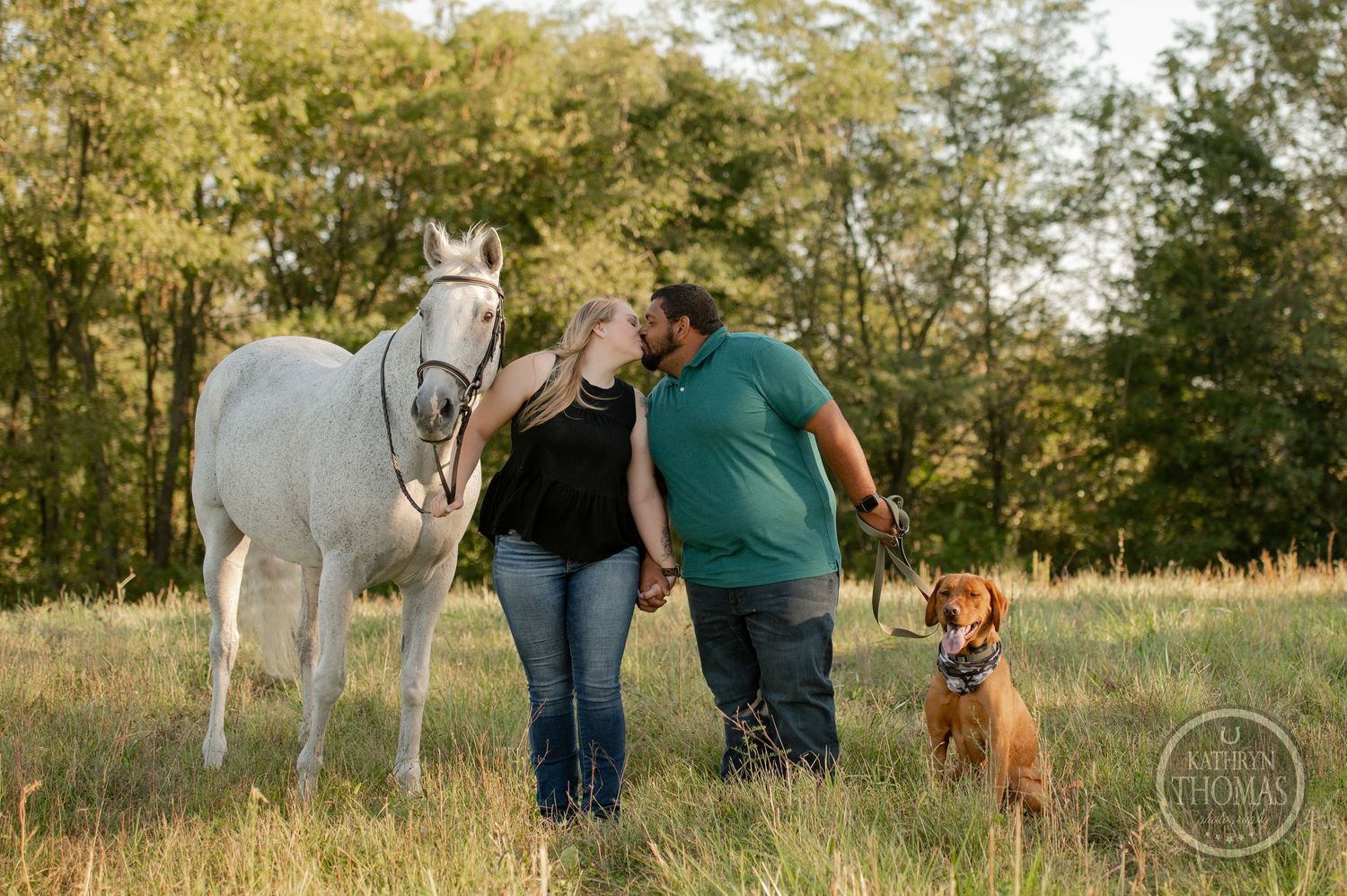 A couple shares a laugh with their white horse and brown dog in a grassy field.