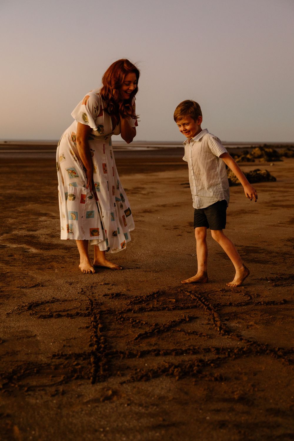 A mother and her son walking on an empty beach at dusk playing tic tac toe.