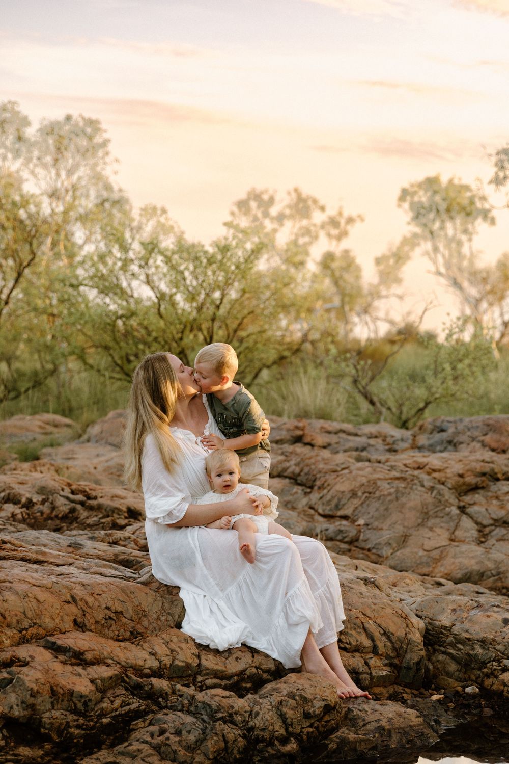 A tender moment at sunset captured on rocky terrain surrounded by lush greenery in warm lighting.